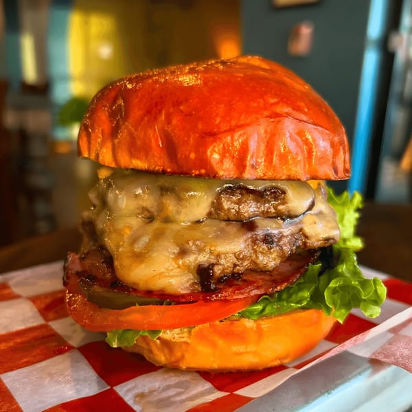 A double patty Franks Burgers cheeseburger with bacon, lettuce, tomato, and pickles on a red and white checkered paper.