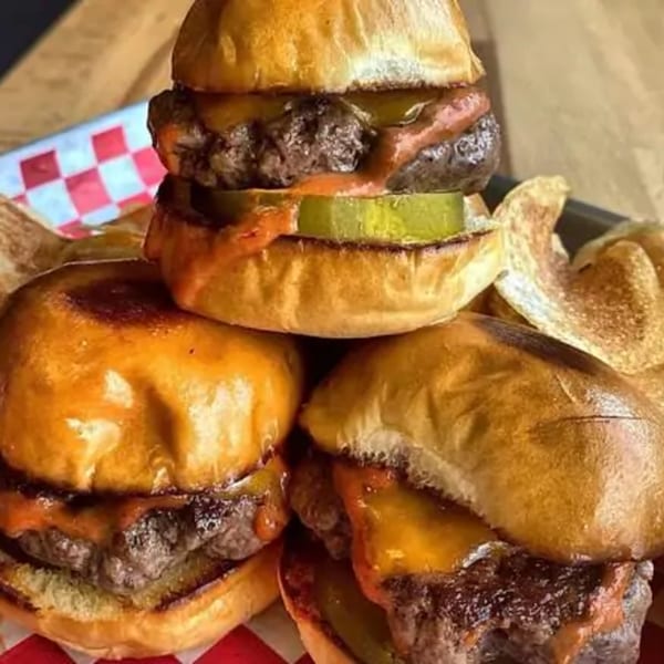 A stack of three Franks Burgers sliders with cheese, sauce, and pickles, served with chips.