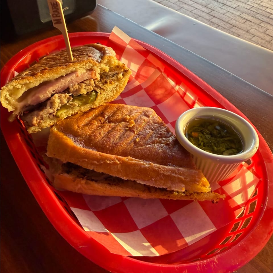 A Frank's Burgers Cuban sandwich, cut in half, with a side of green sauce in a red basket.