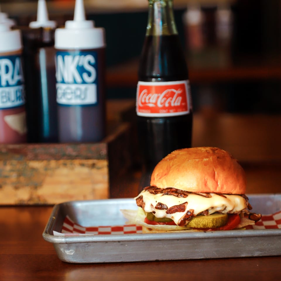 A Franks Burgers cheeseburger with melted cheese, mushrooms, pickles, and tomato, next to a Coca-Cola.