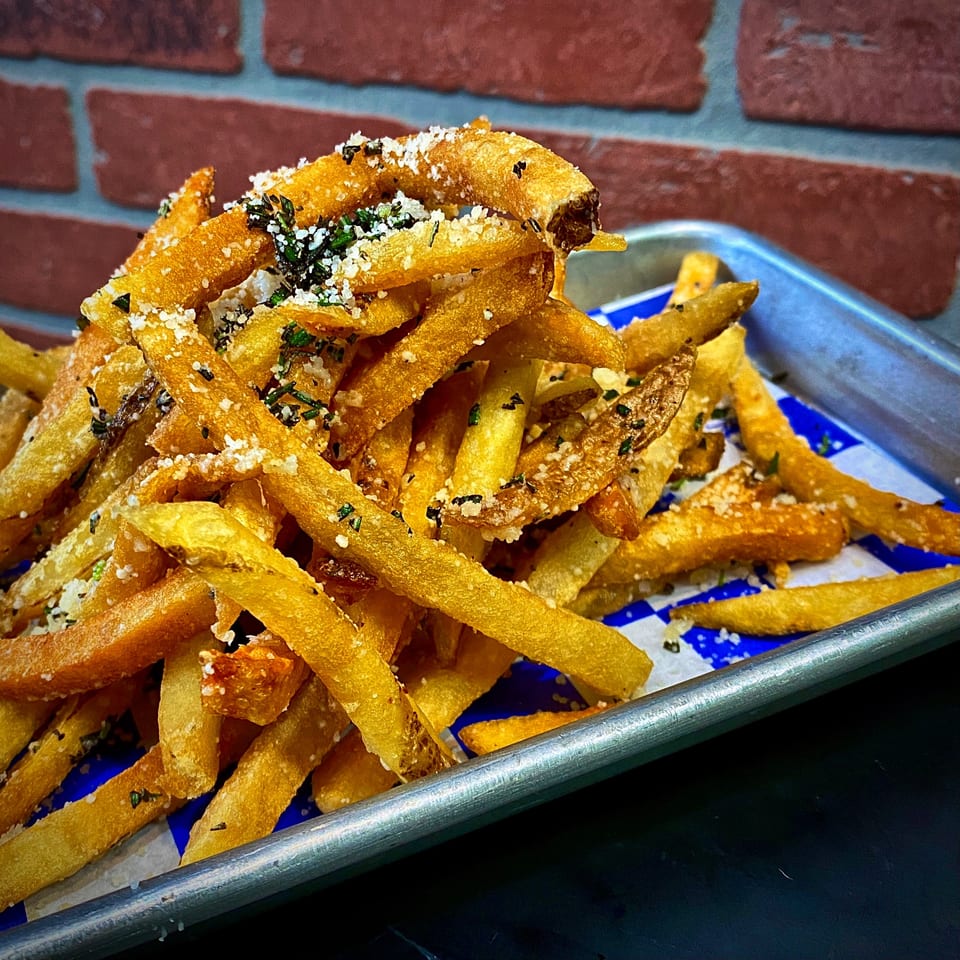 A metal tray filled with golden-brown french fries, topped with parmesan cheese and herbs, against a brick wall.